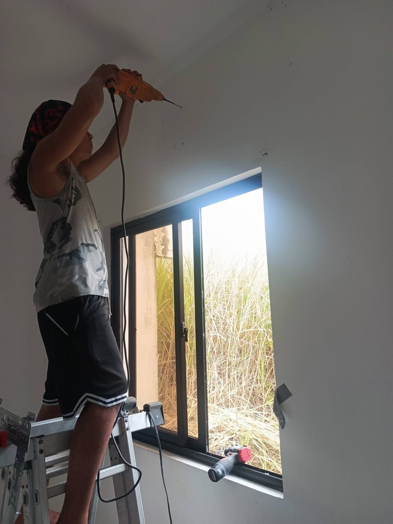Technician drilling the interior wall above a window for a split-type air conditioner installation.