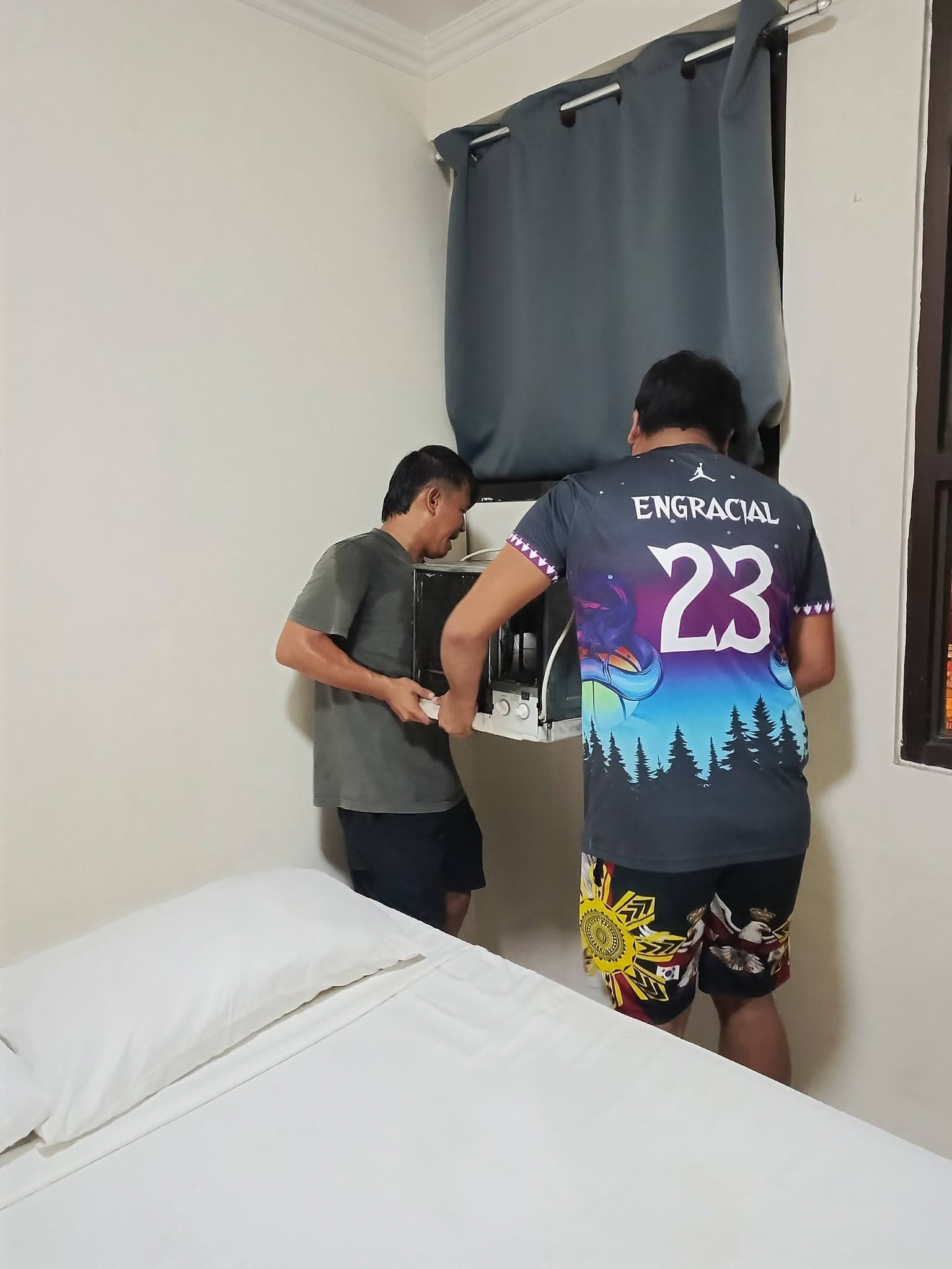 Two technicians lifting a cleaned window air conditioner back into its wall opening.