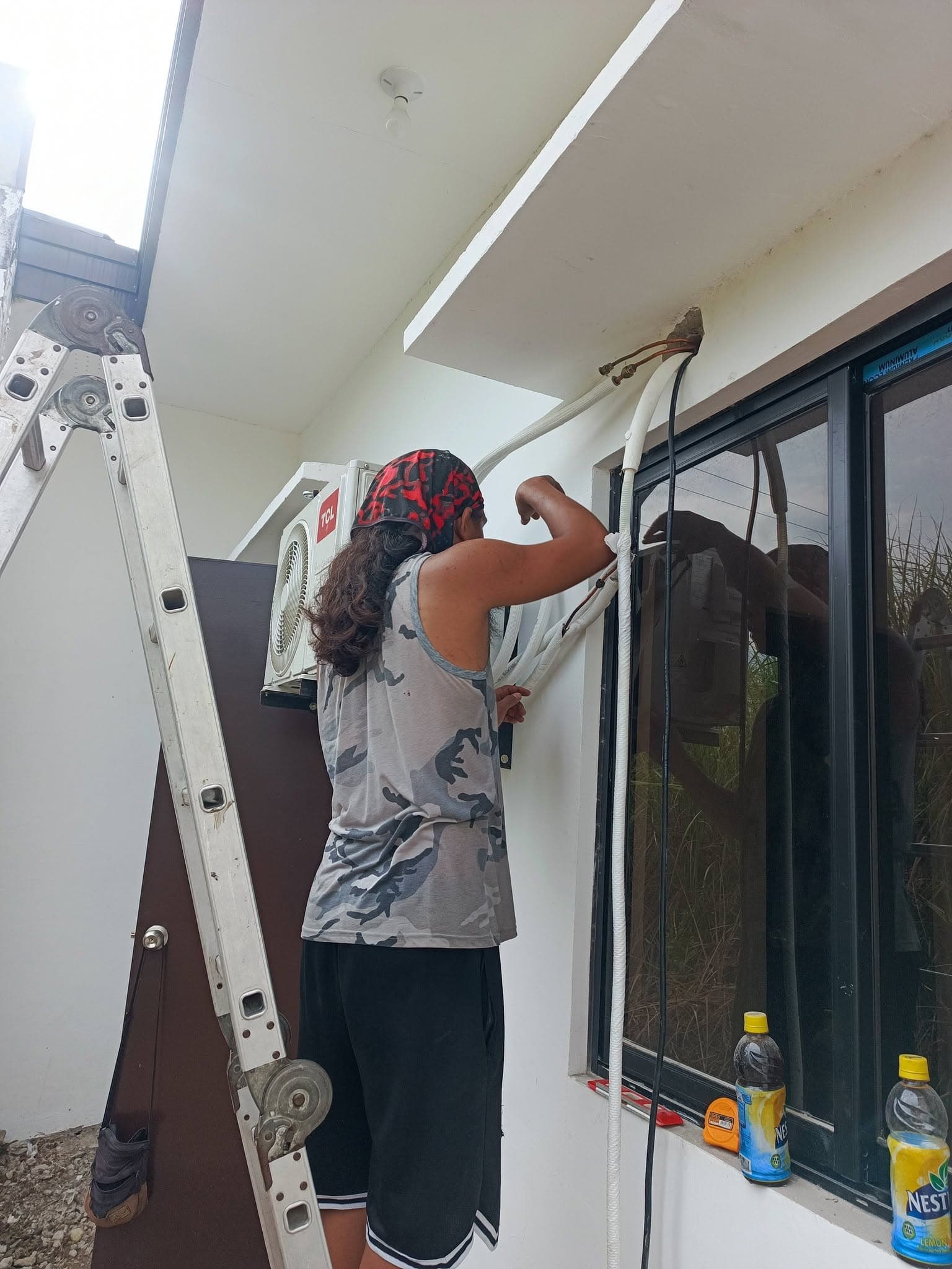 Technician routing insulated refrigerant lines through the exterior wall beside a window.