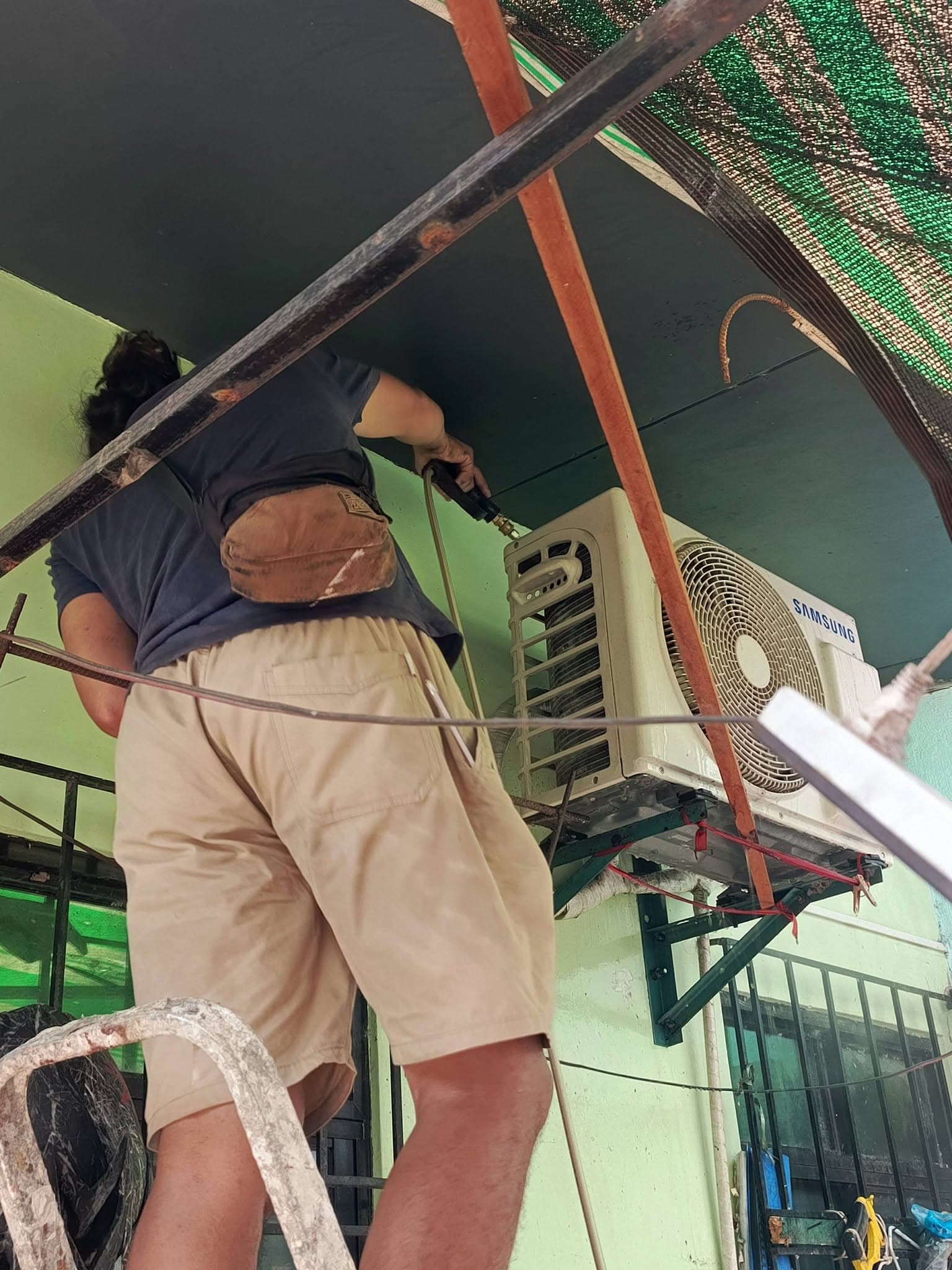 Technician pressure-washing a Samsung outdoor condenser mounted on a wall bracket.