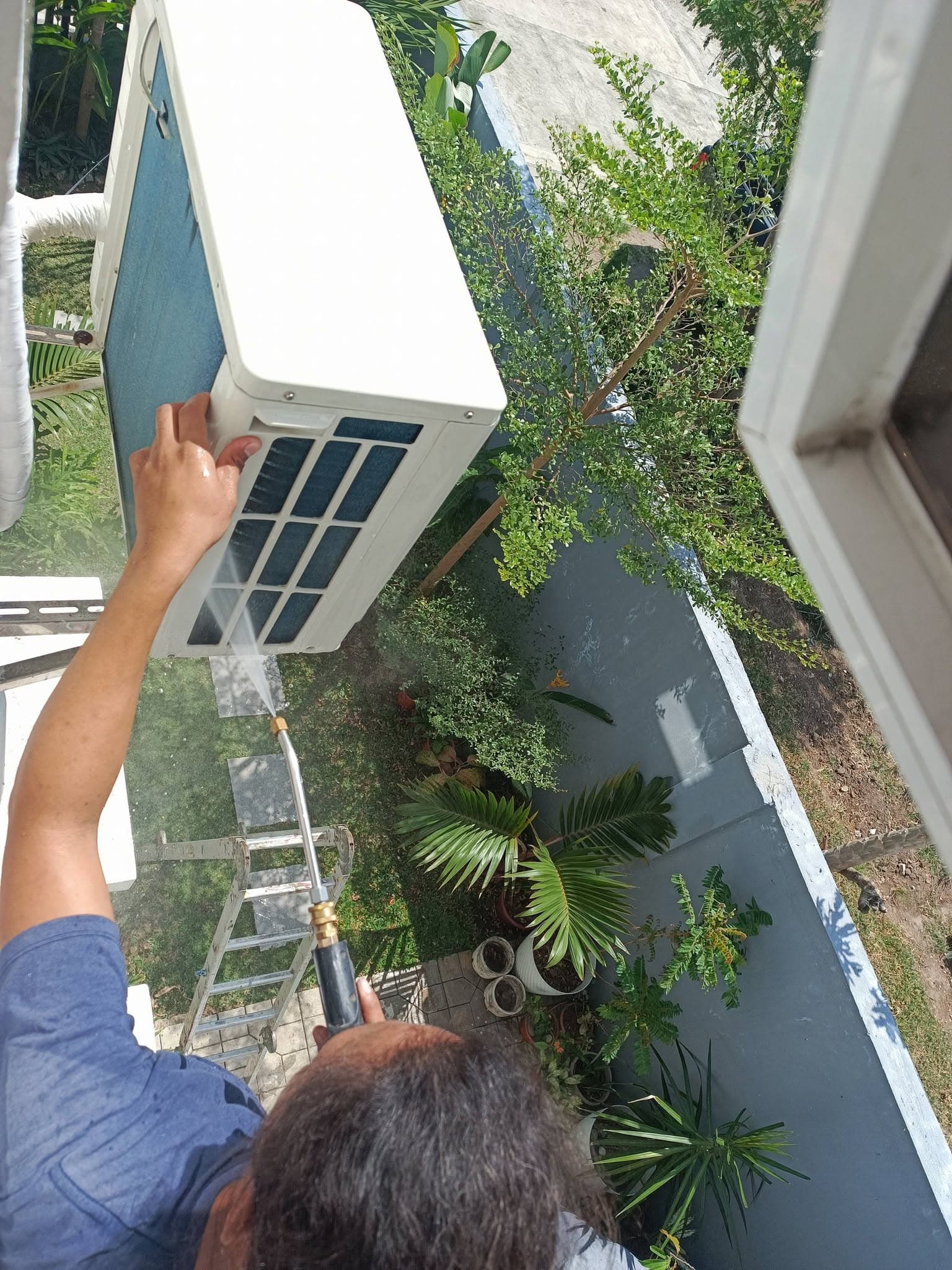 Technician rinsing the underside and coil of an elevated outdoor condenser.