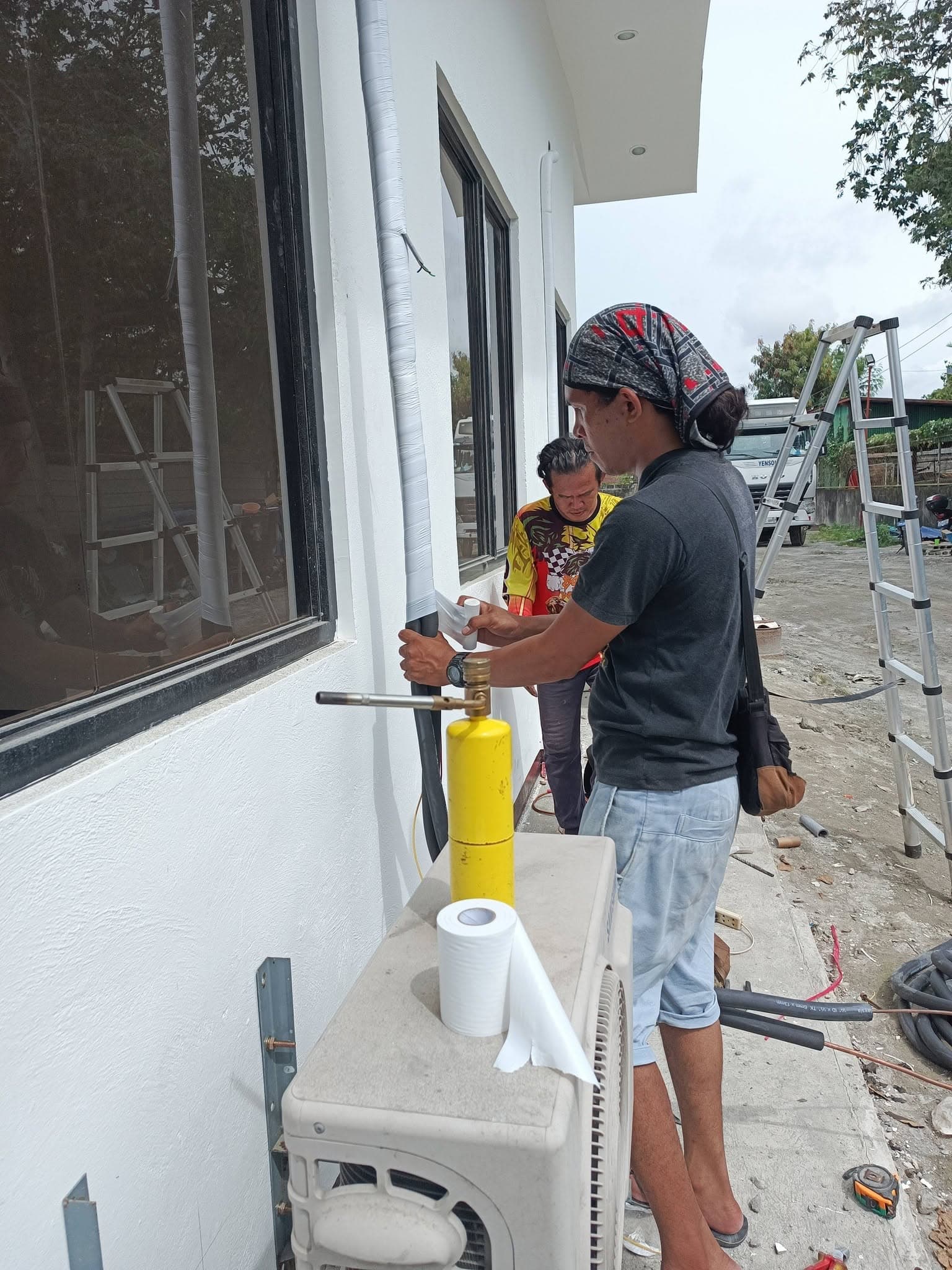 Technicians wrapping and insulating exterior refrigerant lines beside a Samsung condenser.