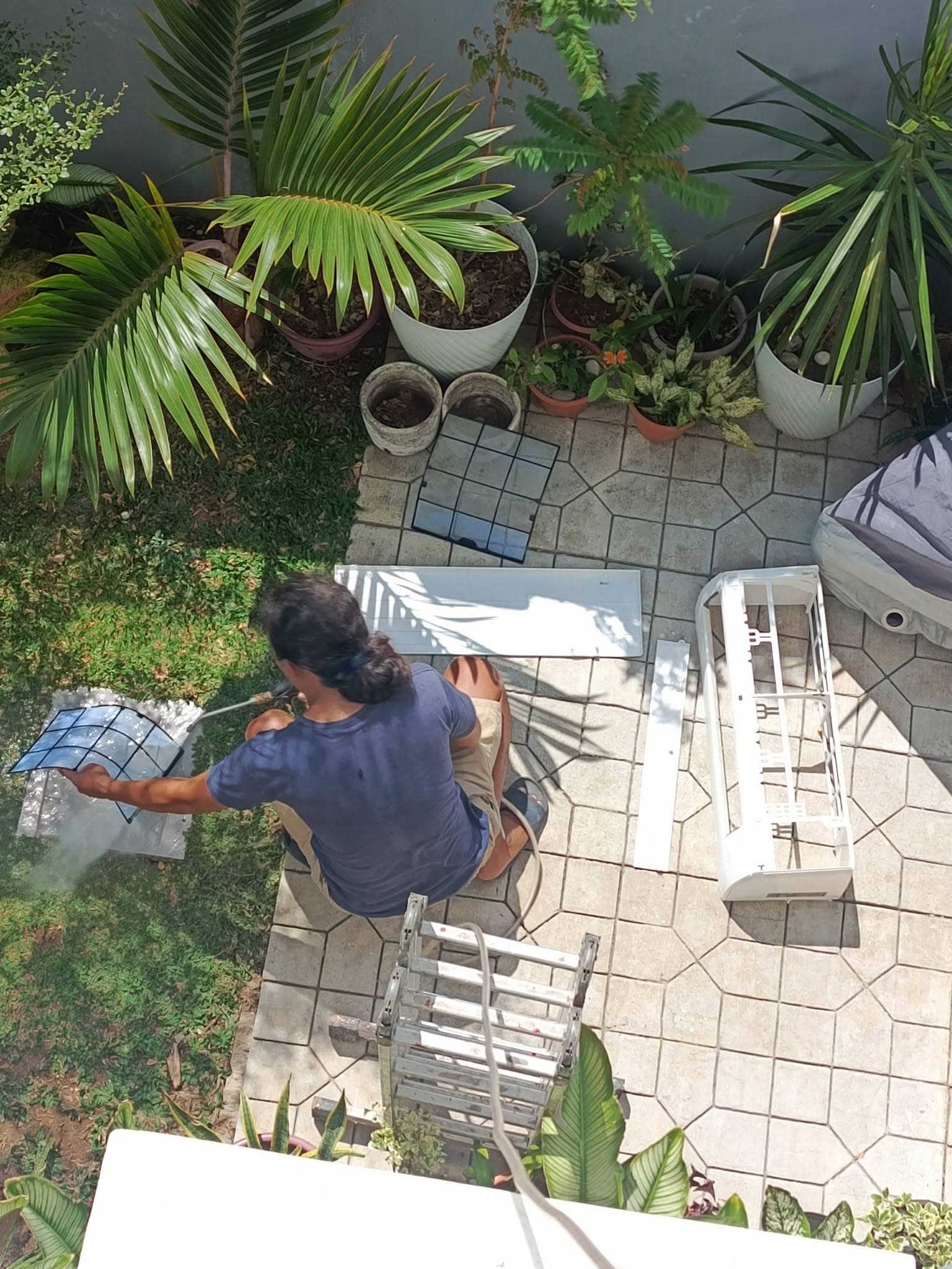 Technician pressure-washing removed filters and indoor-unit panels in a garden work area.